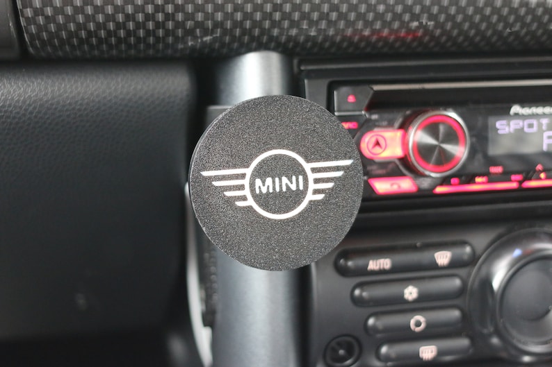 Peut inclure: Un accessoire de voiture rond noir avec le mot "MINI" en lettres blanches et un logo ail&eacute; argent&eacute;.