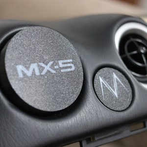 May include: A car dashboard with custom circular accents. The black dashboard has a textured finish. One accent displays "MX-5" and another a stylized "M" logo, both in white. A circular air vent is also visible.