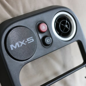 May include: Black plastic car dashboard panel with a round vent, two buttons, and a round emblem that says "MX-5".