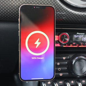 Peut inclure: Un iPhone dor&eacute; avec une ic&ocirc;ne de charge blanche sur un &eacute;cran d&eacute;grad&eacute; violet et rouge. Le t&eacute;l&eacute;phone est mont&eacute; dans une voiture et affiche "100% charg&eacute;".