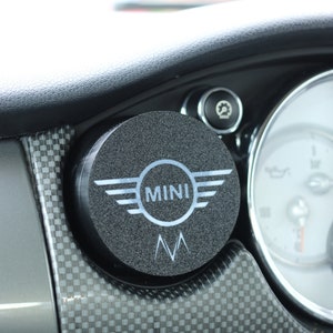 Peut inclure: Un accessoire de voiture rond noir avec le mot "MINI" en lettres blanches et un design d'aile stylisé. L'accessoire a une surface texturée et un petit logo "M".