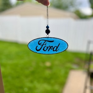 May include: A blue oval-shaped ornament with the word "Ford" in black script. The ornament is suspended by a thin silver string, with a black and blue bead accent. The background is a blurred outdoor scene.