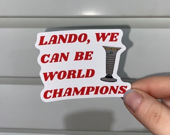 May include: A white sticker with red text that reads "Lando, We Can Be World Champions". A cartoon image of a trophy is next to the text.