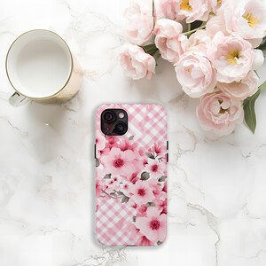 May include: A pink and white gingham patterned phone case with a floral design featuring pink flowers and green leaves. The case is next to a gold mug and a bouquet of pink flowers.