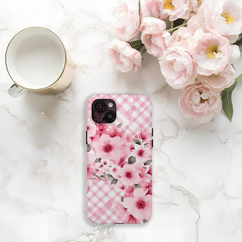 May include: A pink and white gingham patterned phone case with a floral design featuring pink flowers and green leaves. The case is next to a gold mug and a bouquet of pink flowers.