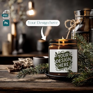 Op de afbeelding: Een brandende kaars in een bruine glazen pot met een wit etiket met de tekst "YOUR DESIGN HERE" in het groen. De kaars staat op een zwarte schotel, omringd door dennentakken, op een houten oppervlak. De achtergrond bevat andere decoratieve items.