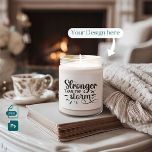 May include: A lit, white candle in a glass jar with the text "Stronger Than The Storm" in black script. The candle sits on a stack of books, with a teacup and a cozy blanket in the background. The words "Your Design here" are also visible.
