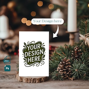 Op de afbeelding: Een witte kaart met de tekst "YOUR DESIGN HERE" in een groen, decoratief ontwerp, weergegeven op een houten basis. De afbeelding bevat een JPEG- en Ps-pictogram, met een vage achtergrond van kerstversieringen en lichten.