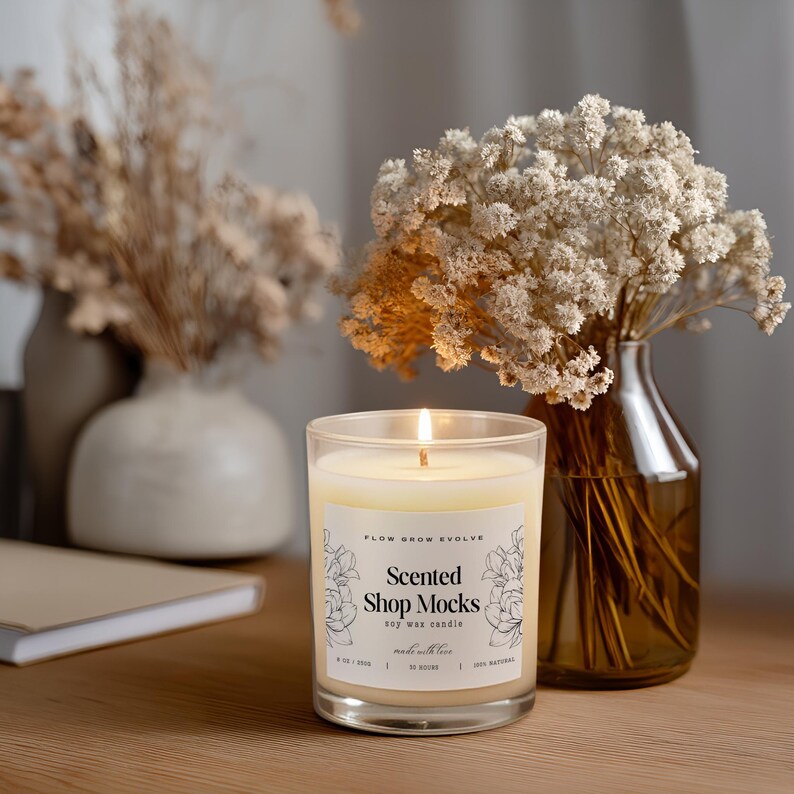 May include: A lit soy wax candle in a clear glass jar with a label that reads "Scented Shop Mocks". The candle is next to a vase of white flowers and a notebook. The candle is on a wooden surface.