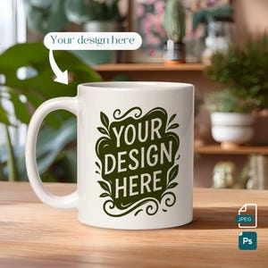May include: A white ceramic mug with a large handle. The mug features the text "YOUR DESIGN HERE" in a dark green, stylized font, surrounded by decorative leaves and swirls. The mug is on a wooden surface.