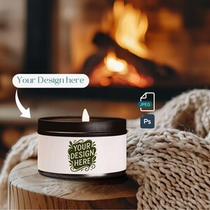 May include: A lit candle in a black metal container with a white label. The label features the text "Your Design Here" in a green, decorative font. The background includes a cozy fireplace and a knitted blanket.