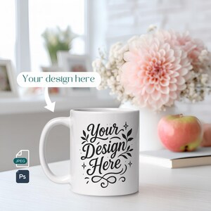 Può includere: Tazza in ceramica bianca con la scritta "Your Design Here" in un carattere decorativo. La tazza è su una superficie bianca, con un bouquet di fiori rosa e una mela sullo sfondo. L'immagine include anche un'icona JPEG e Ps.