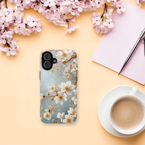 May include: A smartphone case featuring a floral design with white flowers and gold accents on a light blue background. The case is surrounded by pink flowers, a cup of coffee, a pen, and a notepad on a light yellow surface.