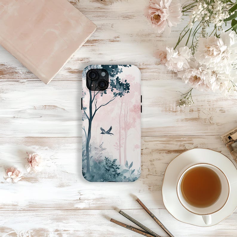 May include: A white phone case featuring a watercolor forest scene with pink and blue hues. The design includes trees, foliage, and a flying bird. The case is on a white wooden surface, with flowers and a cup of tea.