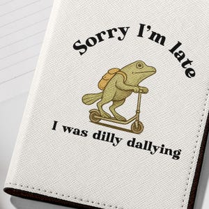 May include: White passport holder with a frog riding a scooter and the text "Sorry I'm late I was dilly dallying".
