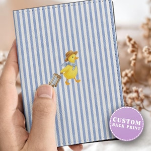 May include: Passport holder with a blue and white striped pattern. The cover features an illustration of a yellow duck wearing a hat and scarf, pulling a suitcase. The holder has a custom back print option.