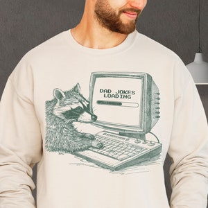 May include: A beige sweatshirt with a graphic of a raccoon using a computer. The computer screen displays the text "Dad Jokes Loading" with a progress bar.
