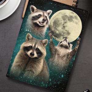 May include: A black passport holder featuring a whimsical illustration of three raccoons interacting with a full moon against a starry teal background. The passport holder has a brown spiral notebook and a cup of coffee in the background.