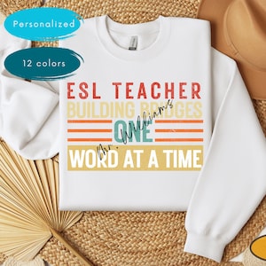 May include: White sweatshirt with a vintage-style graphic that says "ESL Teacher Building Bridges One Word At A Time" with the name "Williams" added. The text is in red, blue, and yellow.