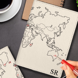 May include: Cream-colored passport holder with a black world map design and red marker accents. The passport holder has the initials "SR" and is next to a cup of coffee and a pen. The image is of a travel-themed product.