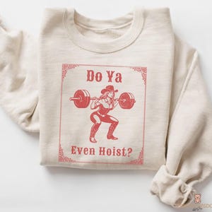 May include: A beige sweatshirt featuring a red graphic of a woman in a cowboy hat lifting a barbell. The text "Do Ya Even Hoist?" is displayed in a decorative frame. The sweatshirt is folded.