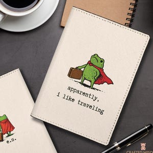 May include: Off-white passport holder with a cartoon frog wearing a red cape and carrying a suitcase. The text "apparently, I like traveling" is printed below the frog. A second passport holder is partially visible.