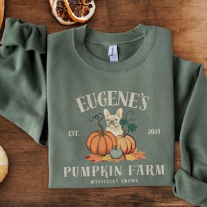 May include: A green sweatshirt with a white graphic design that says "Eugene's Pumpkin Farm Est. 2024 Magically Grown". The graphic also features a cartoon French bulldog wearing a bow tie and sitting on top of pumpkins.