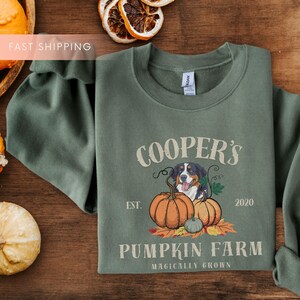 May include: A green sweatshirt with a white graphic design that says "COOPER'S EST. 2020 PUMPKIN FARM MAGICALLY GROWN". The graphic also features a Bernese Mountain Dog and pumpkins.