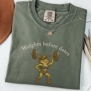 May include: Olive green t-shirt with the phrase "Weights before dates" and a cartoon frog lifting weights. The frog is wearing a cowboy hat, boots, and a crop top. A silver necklace and a brown hat are also visible.