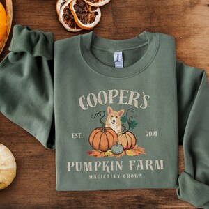 May include: A green sweatshirt with a white graphic design that says "Cooper's Pumpkin Farm Established 2021 Magically Grown". The graphic also features a cartoon corgi dog sitting on top of pumpkins.