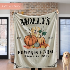 May include: Cream-colored blanket featuring a golden retriever, pumpkins, and the text "MOLLY'S PUMPKIN FARM MAGICALY GROWN EST. 2024". The design includes orange and green pumpkins with fall leaves. The blanket has decorative border details.