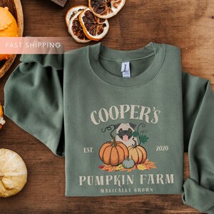 May include: Sage green sweatshirt featuring a pug dog illustration with pumpkins and the text "COOPER'S PUMPKIN FARM". The design includes the year "2020" and the phrase "Magically Grown". A cozy fall-themed apparel item.