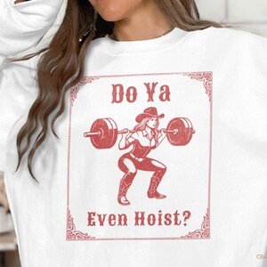 May include: White sweatshirt with a red graphic of a woman in a cowboy hat lifting weights. The text "Do Ya Even Hoist?" is displayed in a decorative frame. The design is a vintage-style illustration.