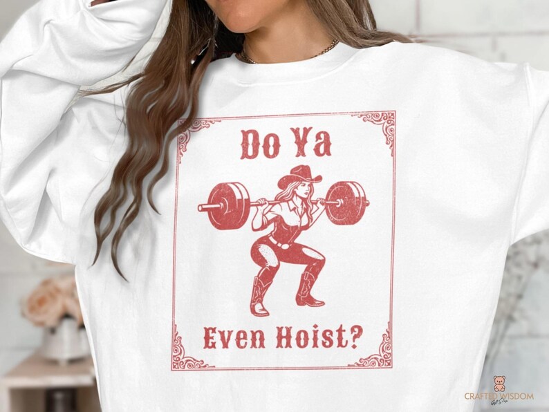 May include: White sweatshirt with a red graphic of a woman in a cowboy hat lifting weights. The text "Do Ya Even Hoist?" is displayed in a decorative frame. The design is a vintage-style illustration.