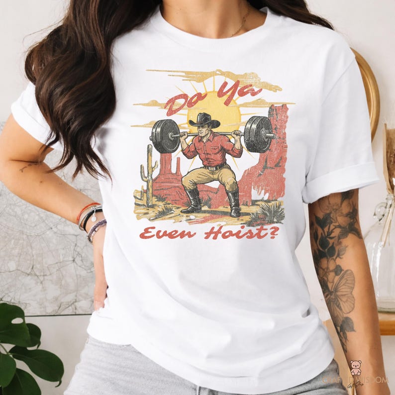 May include: White t-shirt featuring a graphic of a cowboy lifting weights in a desert landscape. The design includes the text "Do Ya Even Hoist?" in a vintage style, with a sun and cacti in the background.