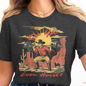 May include: Dark gray t-shirt featuring a vintage-style graphic of a cowboy lifting weights in a desert landscape. The design includes the text "Do Ya Even Hoist?" in a distressed font, with a sun and cacti.