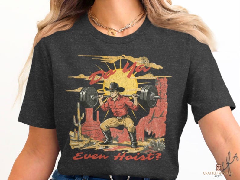May include: Dark gray t-shirt featuring a vintage-style graphic of a cowboy lifting weights in a desert landscape. The design includes the text "Do Ya Even Hoist?" in a distressed font, with a sun and cacti.