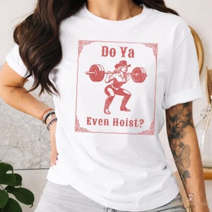 May include: White t-shirt with a red graphic of a cowgirl lifting weights, framed by a decorative border. The text "Do Ya Even Hoist?" is above and below the illustration. The shirt has short sleeves.