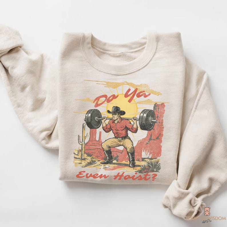 May include: Cream-colored sweatshirt featuring a graphic of a cowboy lifting weights in a desert landscape. The design includes a cowboy in a red shirt and black hat, lifting a barbell. Text reads "Do Ya Even Hoist?"