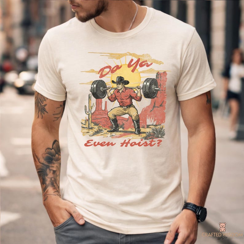 May include: Cream-colored t-shirt featuring a graphic of a cowboy lifting weights in a desert landscape. The design includes the text "Do Ya Even Hoist?" in red and yellow hues, with a cartoon-style illustration.