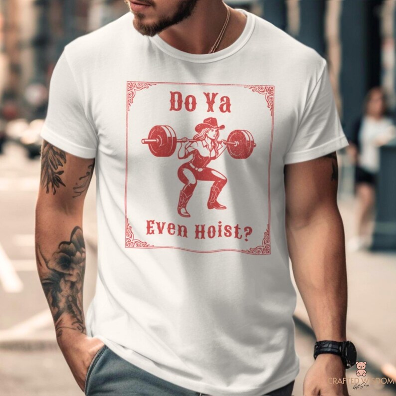 May include: White t-shirt featuring a red graphic of a cowgirl lifting weights with the text "Do Ya Even Hoist?" in a decorative frame. The design includes a cowgirl in a squat position, wearing a hat and boots.
