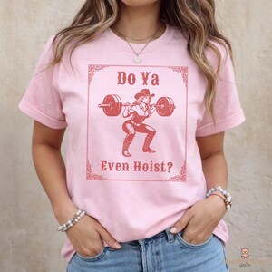 May include: Pink t-shirt with a graphic of a cowgirl lifting weights, the text "Do Ya Even Hoist?" is displayed in a decorative frame. The cowgirl is wearing a cowboy hat and boots. The shirt is a short-sleeved crew neck.