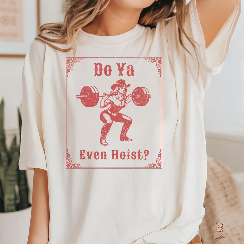 May include: Cream-colored t-shirt with a red graphic of a cowgirl lifting weights, with the text "Do Ya Even Hoist?" in a decorative frame. The design features a cowgirl squatting with a barbell.