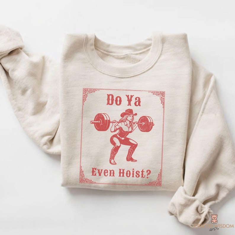 May include: A beige sweatshirt featuring a red graphic of a woman in a cowboy hat lifting a barbell. The text "Do Ya Even Hoist?" is displayed in a decorative frame. The sweatshirt is folded.