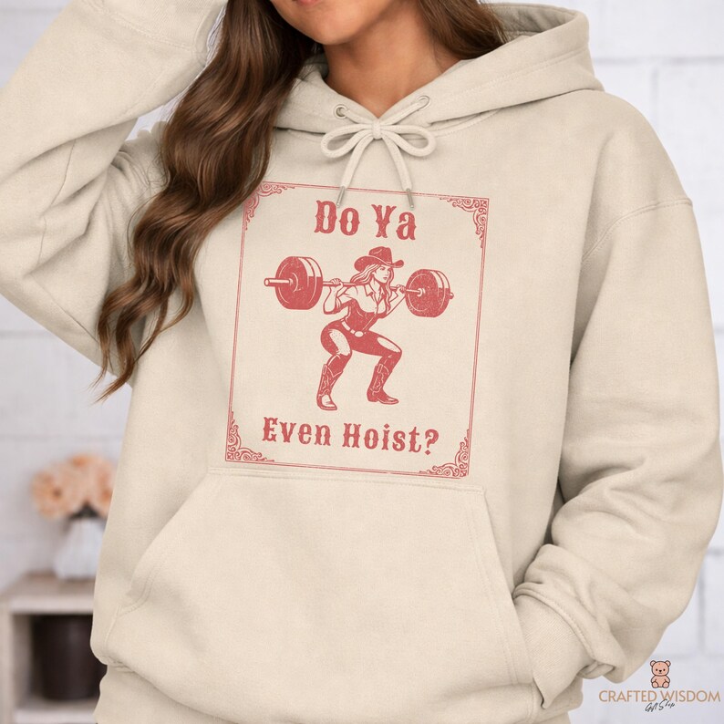 May include: A cream-colored hoodie featuring a red graphic of a woman in a cowboy hat lifting weights. The text "Do Ya Even Hoist?" is displayed above and below the graphic in a decorative frame. The hoodie has a drawstring hood and a front pocket.