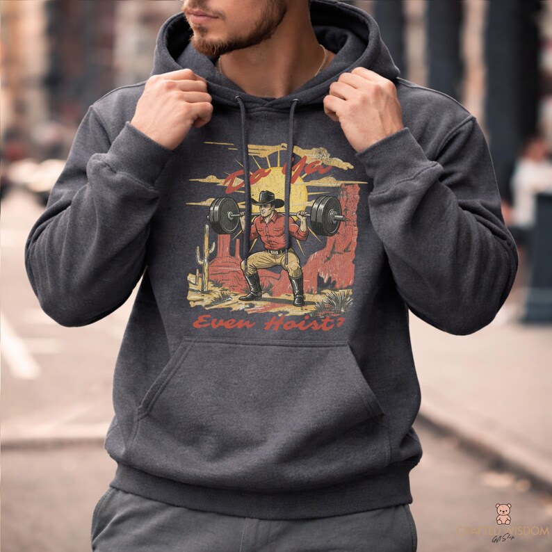 May include: Dark gray hoodie featuring a graphic of a cowboy lifting weights in a desert landscape with a setting sun. The graphic includes the text "Even Hoist?" in red. The hoodie has a front pocket and a drawstring hood.