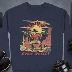 May include: Navy blue sweatshirt featuring a weightlifting cowboy graphic. The design includes a cowboy lifting a barbell with the text "Do You Even Hoist?" in a vintage style. The sweatshirt is surrounded by workout equipment.