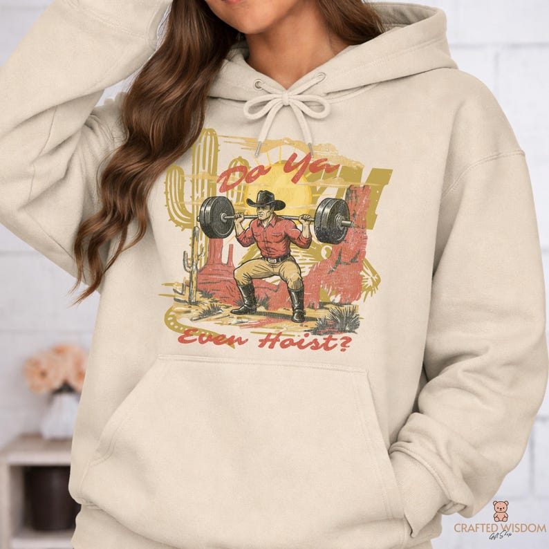 May include: Cream-colored hoodie featuring a graphic of a cowboy lifting weights in a desert scene. The design includes the text "Do Ya Even Hoist?" in a stylized font, with a cactus and red rock formations.