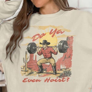 May include: Cream-colored sweatshirt with a graphic of a cowboy lifting weights in a desert scene. The image includes the text "Do Ya Even Hoist?" in a distressed font, with a sun and cacti in the background.
