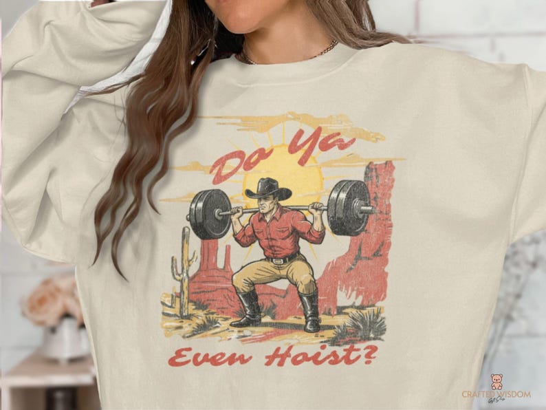 May include: Cream-colored sweatshirt with a graphic of a cowboy lifting weights in a desert scene. The image includes the text "Do Ya Even Hoist?" in a distressed font, with a sun and cacti in the background.